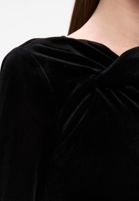 Čierny zamatový top s detailom v podobe skrúteného uzla na výstrihu, hladká štruktúra a jemný lesk, ktorý predvádza draped dizajn.