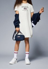 Světle šedé tričkové šaty s potiskem loga, tmavě modrý svetr přehozený, tmavě modrá taška přes rameno, bílé tenisky a bílé ponožky s červenými pruhy.