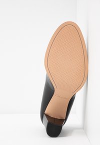 Semelle d'une chaussure en cuir noir avec une semelle extérieure en caoutchouc beige et un petit talon empilé, positionnée à la verticale contre un fond d'angle blanc.