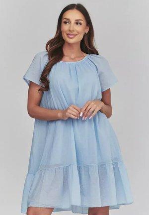 Kobieta z długimi brązowymi włosami, ubrana w luźną, jasno niebieską sukienkę z krótkim rękawem, uśmiechająca się i patrząca lekko na bok na jednolitym tle.