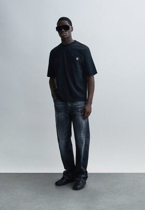 Black short-sleeve T-shirt with a pocket, paired with faded blue straight-leg jeans. Worn with black sneakers and oversized black sunglasses.