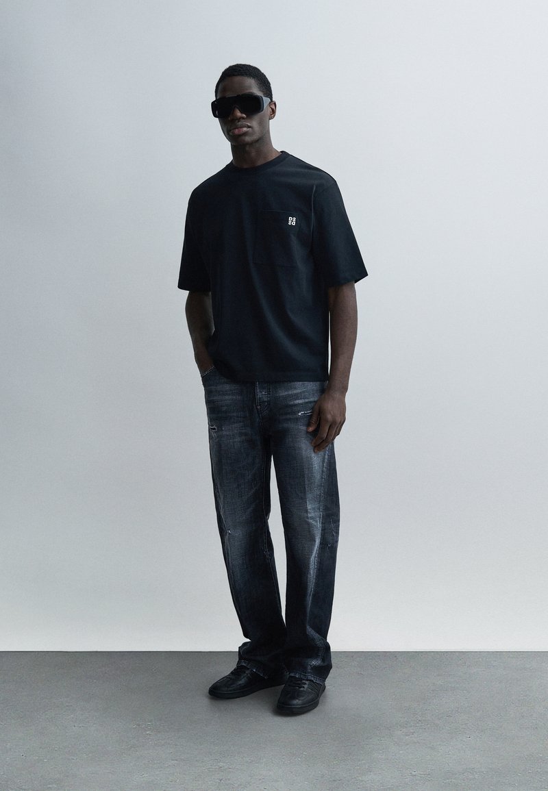 Μαύρο κοντομάνικο T-shirt με τσέπη, συνδυασμένο με ξεβαμμένο μπλε τζιν με ίσια γραμμή. Φορεμένο με μαύρα αθλητικά παπούτσια και μεγάλα μαύρα γυαλιά ηλίου.