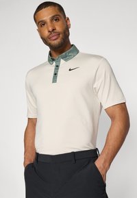 Nike polo krekls krēmkrāsā ar tumši zaļu apkakli, kas aprīkots ar pogu plāksteri un melnu logo. Īsas piedurknes ar gludu auduma tekstūru.