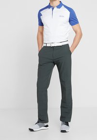 Vit och blå polo med geometriskt mönster, mörkgröna byxor och grå sportskor, kompletterat med ett vitt bälte.