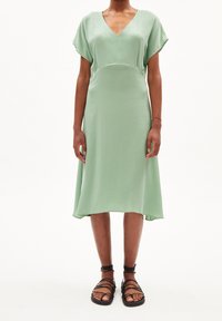 Miętowa sukienka do kolan z dekoltem w kształcie V, krótkimi rękawami i sylwetką w kształcie litery A. Noszona z czarnymi sandałami na cienkich paskach.