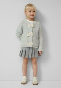 Jasnoszary kardigan o fakturze z trzema kremowymi kokardkami, w zestawie z plisowaną szarym spódnicą i białymi sneakersami. Gładki materiał i luźny krój.