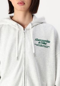 Vaaleanharmaa vetoketjuhuppari, jossa on nyörillä varustettu huppu ja vihreä brodeerausteksti rinnassa. Valmistettu pehmeästä materiaalista, jossa on rento malli.