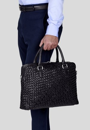 Black leather briefcase with woven texture, dual top handles, silver hardware, and zip closure. Displayed being held against navy trousers.