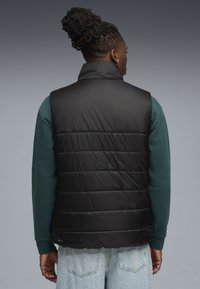 Sort, polstret vest med høj krave og vandret syninger, båret over en grøn sweatshirt, set fra ryggen. Denim jeans er synlige.