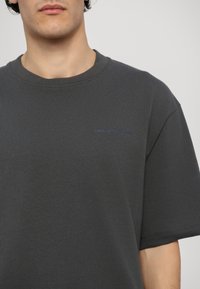 T-shirt en coton gris foncé à manches courtes. Présente un col rond et un logo bleu discret sur la poitrine. Texture lisse. Design simple.