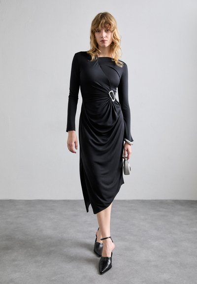 Vestido negro de manga larga con un diseño retorcido, que presenta un detalle plateado. Tela suave, dobladillo asimétrico, combinado con tacones negros de punta.