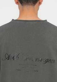Czarnoszary, oversize t-shirt z teksturowanym wykończeniem, z logo i adresem strony internetowej nadrukowanym w kontrastującym, błyszczącym czarnym kolorze na plecach.