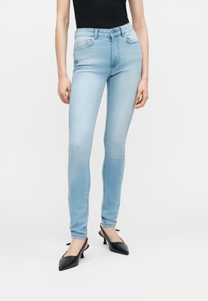 Femme portant un jean skinny bleu clair, un haut noir sans manches et des escarpins noirs à bout pointu avec bride arrière, debout devant un fond uni.