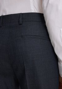 Nahaufnahme einer Person, die dunkle, strukturierte Anzughosen mit einer Gesäßtasche und einem gesteckten weißen Langarmhemd trägt.