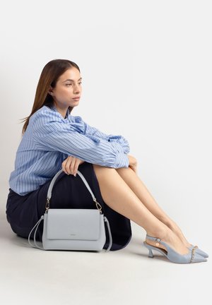 Donna con camicia a righe blu e gonna blu navy seduta con borsa azzurro chiaro e décolleté slingback abbinate su sfondo bianco.