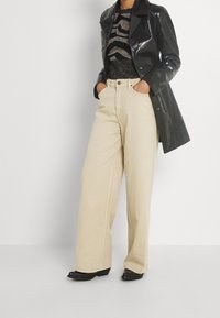 Veste en cuir verni noir, pull en maille noire à motifs scintillants, pantalon beige à jambes larges, et chaussures noires à talons blocs avec ouverture à l'avant.