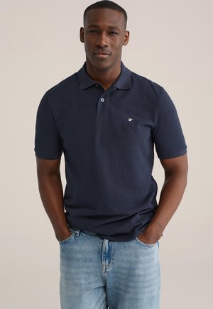 Polo bleu marine en coton à manches courtes, patte de boutonnage à trois boutons et petit logo brodé sur la poitrine gauche. Porté avec un jean bleu clair.