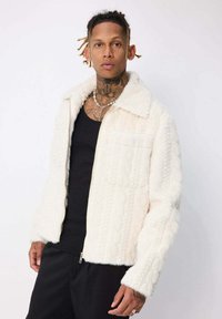 Homme avec des tatouages sur le cou et les mains portant une veste zippée blanche texturée sur une chemise noire et un pantalon noir, sur un fond uni.