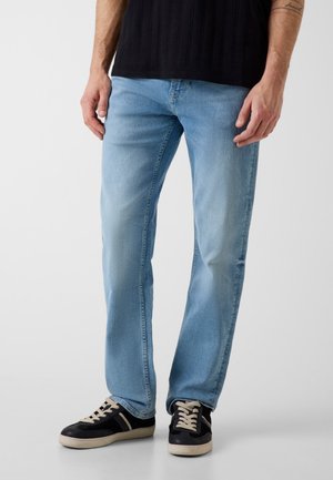 Homme portant un jean bleu clair et des baskets noires avec des lacets blancs, debout devant un fond blanc uni.