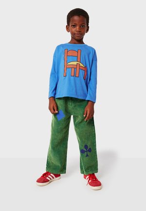 Blaues Langarmshirt mit abstrakter orangefarbener Grafik; grüne Cordhosen mit blauem Patch und Kleeblattmuster; rote Turnschuhe mit weißen Streifen.