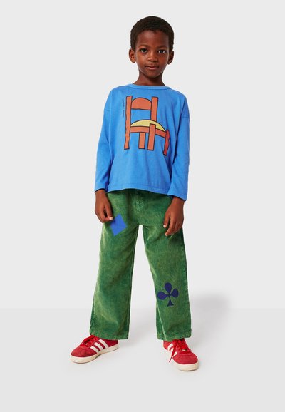 Blå långärmad skjorta med abstrakt orange grafik; gröna kordbyxor med blå lapp och klöverdesign; röda sneakers med vita ränder.