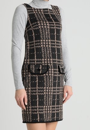 Mouwloze zwart-beige geruite jurk met twee voorzakken met knopen, gedragen over een lichtgrijs truitje met col en lange mouwen.