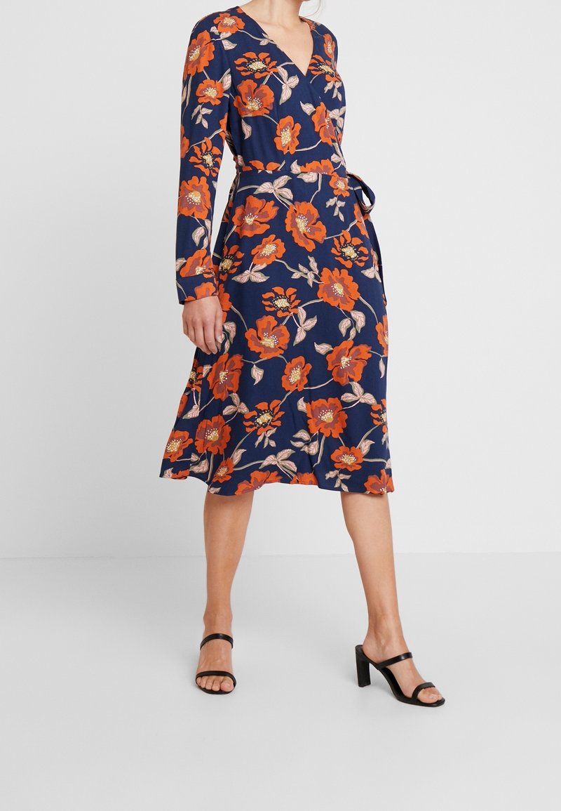 Femme portant une robe portefeuille midi bleu marine avec un grand imprimé floral orange et des sandales noires à talons hauts, debout devant un fond uni.