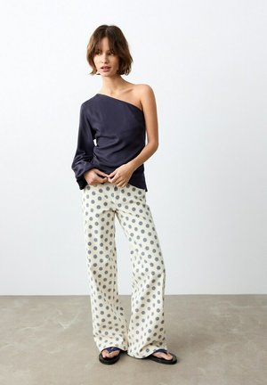 Femme portant un haut asymétrique bleu marine foncé, un pantalon ample crème avec des motifs de soleil bleus, et des sandales noires, debout contre un mur uni.