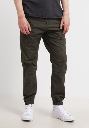 Mann trägt dunkelgrüne Cargo-Jogginghosen und weiße High-Top-Sneaker, steht mit locker an der Seite hängenden Händen.