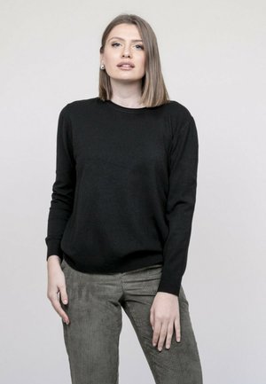 Femme aux cheveux blonds mi-longs portant un pull noir et un pantalon en velours côtelé vert olive, debout devant un fond clair uni.