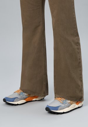 Nogi ubrane w brązowe spodnie o szerokich nogawkach oraz szare, niebieskie i pomarańczowe trampki z beżowymi sznurowadłami na szarym tle.