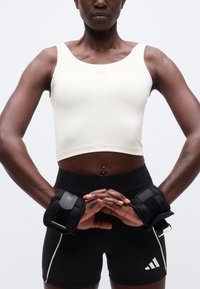 Αθλητικό άτομο που φοράει λευκό crop top και μαύρο σορτς, με τα χέρια μπροστά κλεισμένα, φορώντας μαύρα βάρη καρπού σε κάθε καρπό.