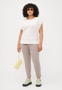 A woman with curly hair stands confidently, wearing a white top with decorative sleeves, beige pants, and green shoes, holding a yellow handbag.