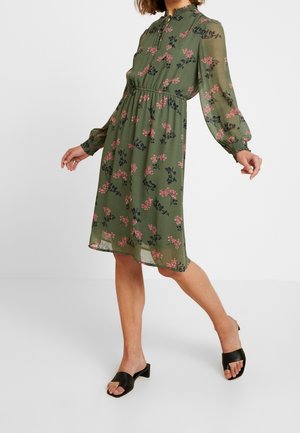 Femme portant une robe verte à fleurs, longueur genou, manches longues avec poignets smockés, associée à des sandales noires à talons blocs et orteils ouverts.