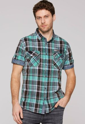 Uomo con camicia a maniche corte a quadri verdi, blu e neri con due tasche sul petto e jeans scuri, in piedi davanti a uno sfondo neutro.