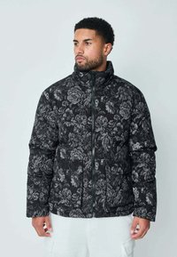 Doudoune noire avec un motif floral dans des nuances de gris. Comprend un col haut, une fermeture éclair à l'avant et des poches latérales. Texture matelassée.