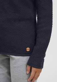 Dark navy knitted sweater with a textured pattern and a wooden logo accent at the bottom. Paired with light gray sweatpants.