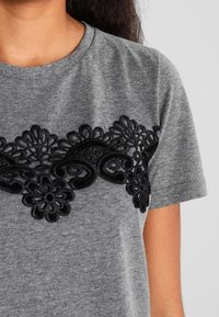 Szara koszulka z krótkim rękawem z czarnymi haftowanymi kwiatowymi detalami na klatce piersiowej, okrągłym dekoltem i miękką teksturą.