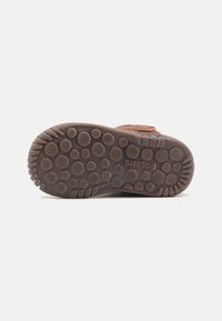 Semelle en cuir marron avec une surface en caoutchouc texturé, présentant des motifs circulaires et des stries pour une meilleure adhérence. Nom de la marque estampé en dessous.