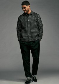 Black patterned shirt with geometrical print, button-up front, and collar. Worn with dark trousers and athletic shoes.