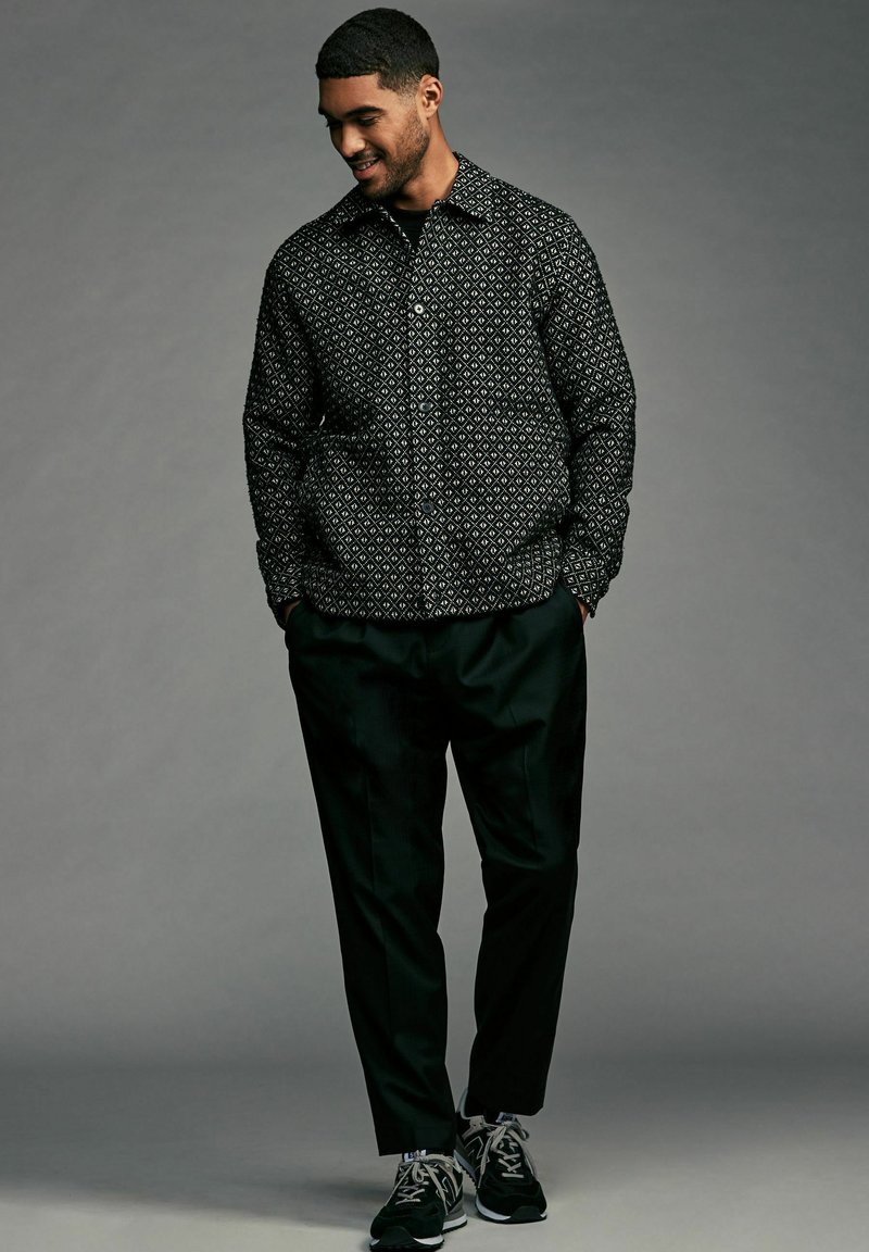 Black patterned shirt with geometrical print, button-up front, and collar. Worn with dark trousers and athletic shoes.