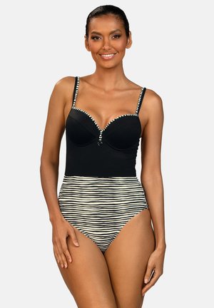 Kobieta w czarnym jednoczęściowym kostiumie kąpielowym z paskami na górze i czarno-białym, poziomo paskowanym dołem, uśmiechająca się z rozluźnionymi rękami.
