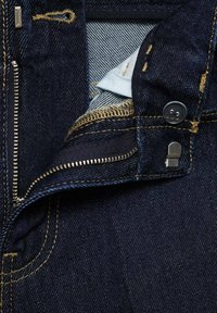 Tmavě modré džíny z denimu s kovovým zipem, zapínáním na knoflík a žlutým prošíváním. Interiér ukazuje detail z látky v světle modré barvě.