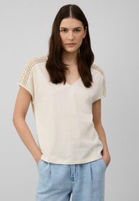 T-shirt crème à col en V avec des manches courtes, pr�ésentant des détails en crochet aux épaules, fabriqué en tissu de coton doux avec une coupe décontractée.