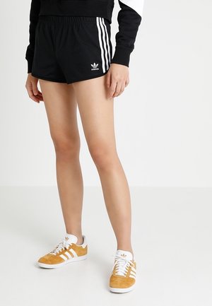 Person iført sorte Adidas-shorts med hvide striber og gule Adidas-sneakers med hvide snørebånd og såler står på en hvid gulv.