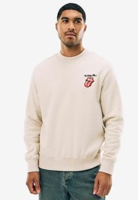 Beige sweatshirt med rund hals, med et broderet rødt læberdesign og teksten "The Rolling Stones". Blødt, tekstureret stof.