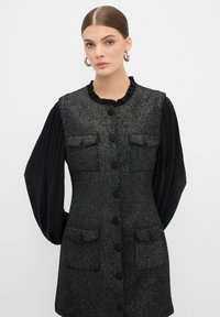 Chaleco negro texturizado con cuello con volantes, cierre frontal de botón y dos bolsillos en el pecho; superpuesto a una blusa negra con mangas plisadas.