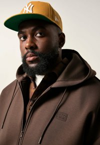 Ein bärtiger Mann trägt einen braunen Zip-Hoodie über einem karierten Hemd und einer senfgelben New York Yankees-Kappe und schaut leicht zur Seite.