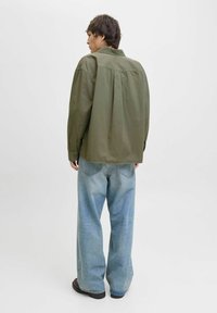 Personne se tenant de profil vers la gauche, portant une chemise oversize vert olive, un pantalon en jean large bleu clair et des chaussures sombres sur un fond uni.