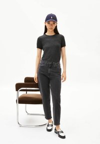 T-shirt noir, jean gris foncé taille haute, mocassins noirs et blancs, et casquette bleu marine avec un accent blanc, debout près d'une chaise marron.